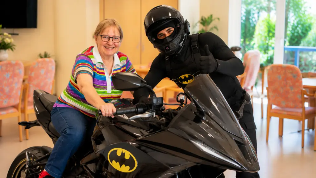 Gästefürsprecherin der anbosa Homecare Tagespflege Palisadenstraße sitzt auf dem Batman-Motorrad von Internetstar Bermo_fit und lächelt begeistert während seines Besuchs in Berlin-Friedrichshain.