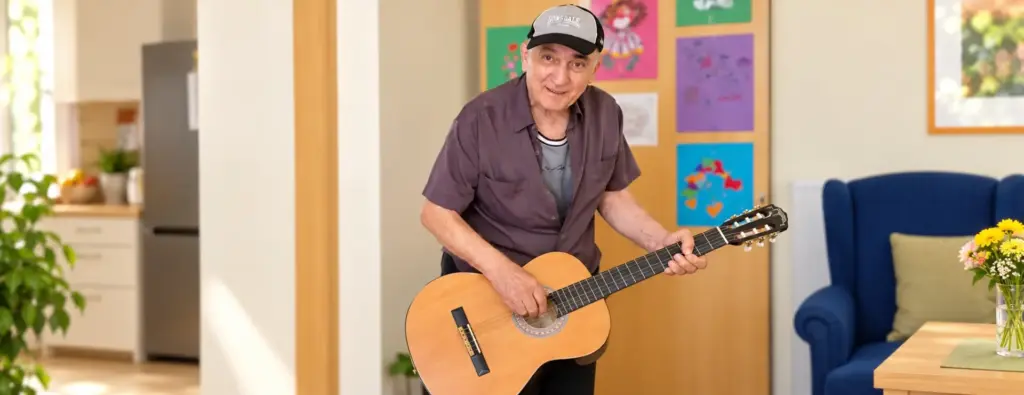 Ein älterer Gast der anbosa Tagespflege Palisadenstraße steht mit einer akustischen Gitarre in einem hellen Gemeinschaftsraum.